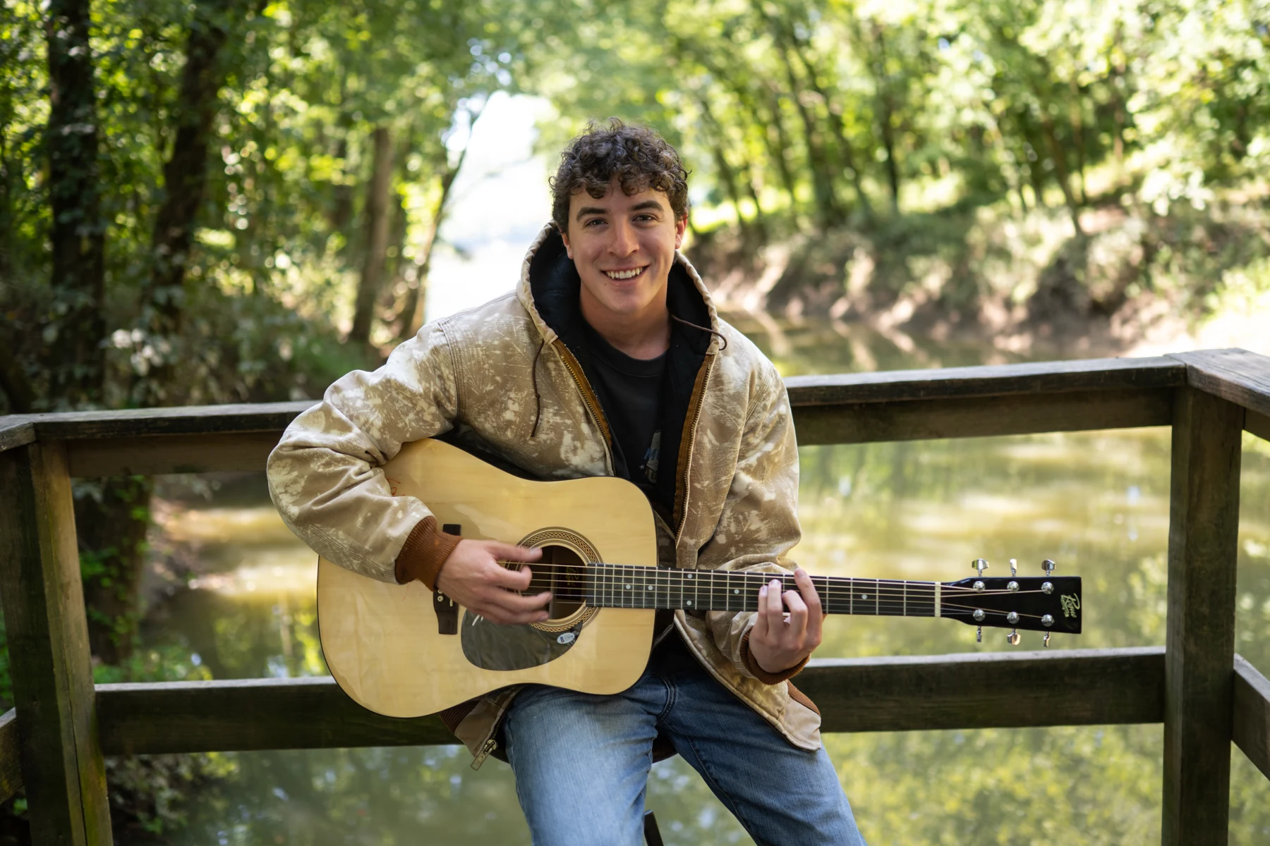 Ryan McKenzie smiling with his acoustic guitar beside a wooded river